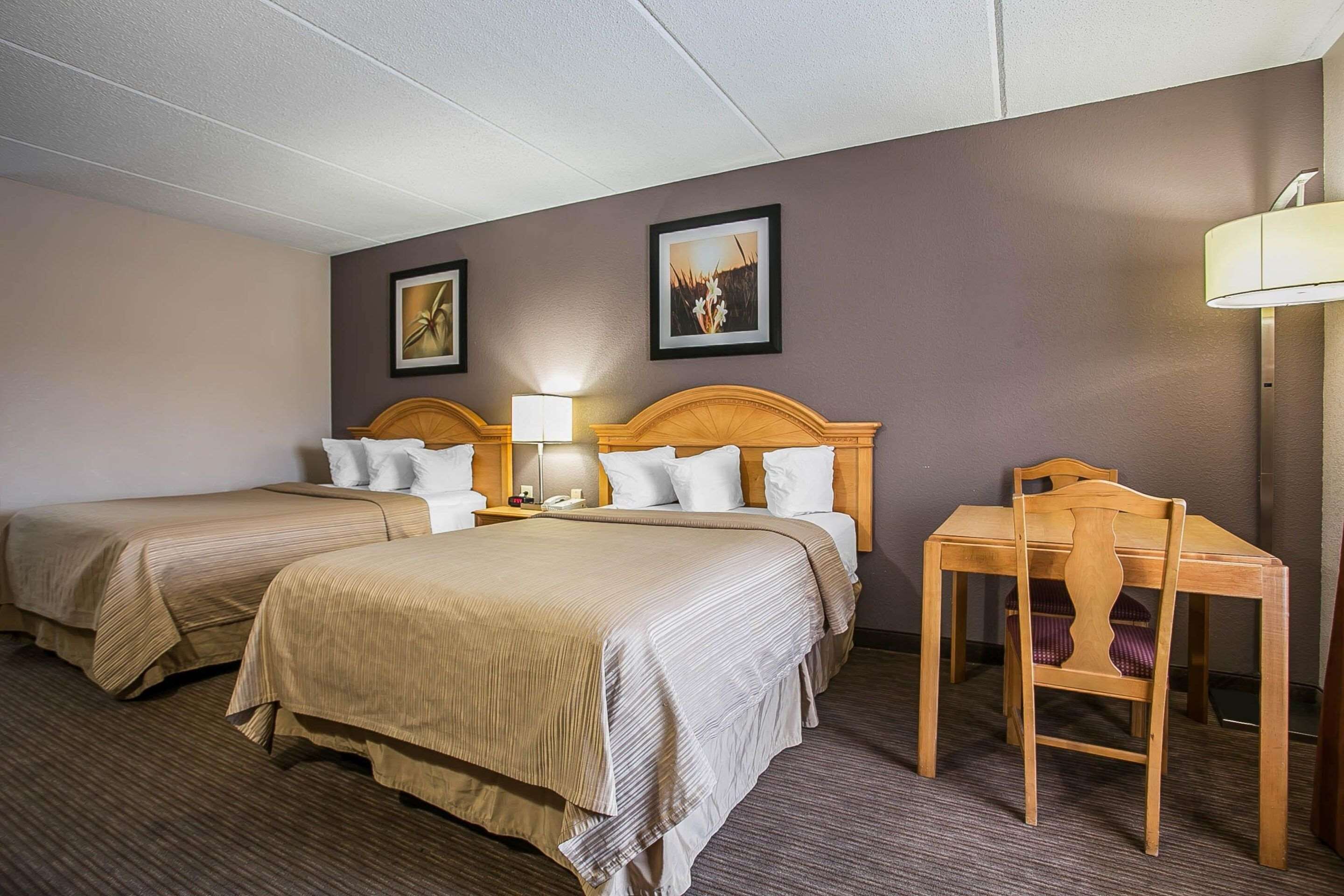 A hotel room with two beds, a desk, and a chair. There are two bedside tables with lamps and a wall-mounted phone.