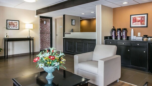 A hotel lobby with a reception desk, a white armchair, a vase of flowers, a black table, a brick wall, and a kitchen area with appliances. - Quality Inn Hartsville (Hartsville)