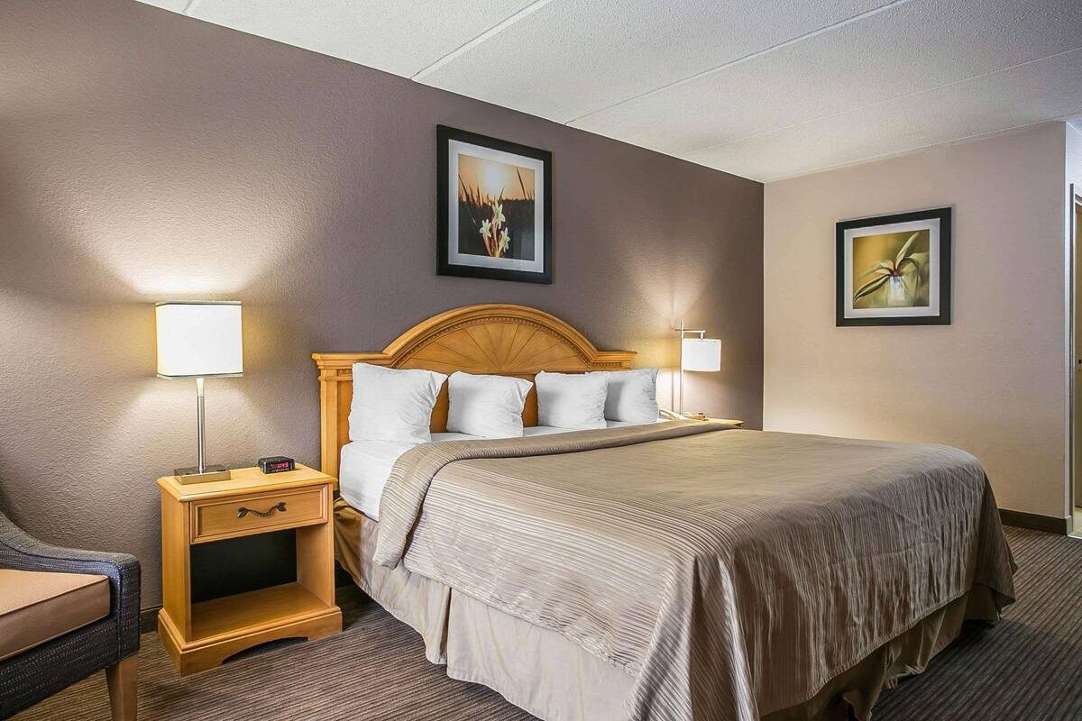 Ein Hotelzimmer mit einem Bett mit Holz-Kopfteil, zwei Nachttischen mit Lampen, einer kleinen Schublade und zwei gerahmten Bildern an den Wänden.