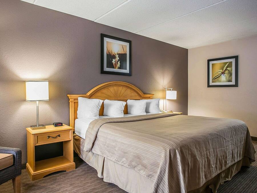 A hotel room with a wooden headboard bed, two bedside tables with lamps, a small drawer, and two framed pictures on the walls.