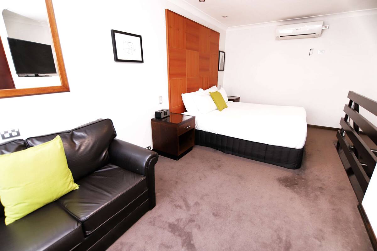 A hotel room with a bed, a wooden headboard, a brown leather sofa with a yellow pillow, a small table with a radio, and a wall-mounted air conditioner.