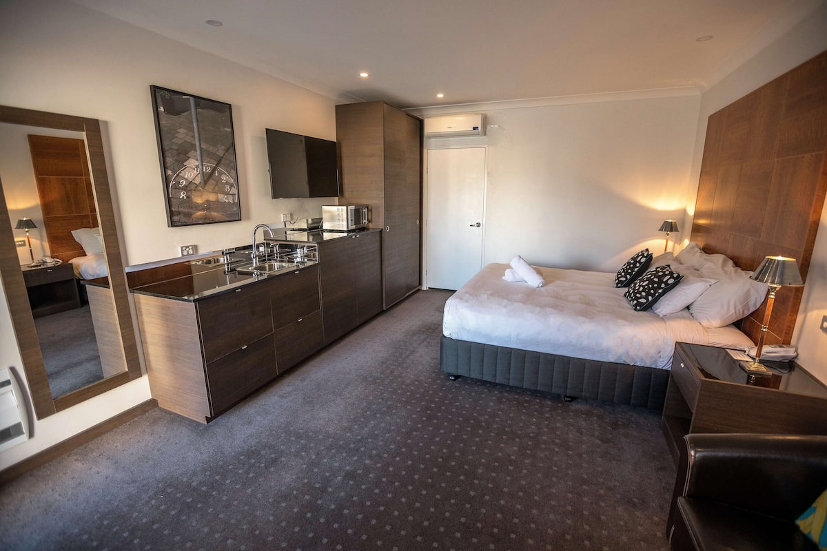 A modern hotel room with a bed, bedside table, lamp, and a built-in kitchen area with a microwave.