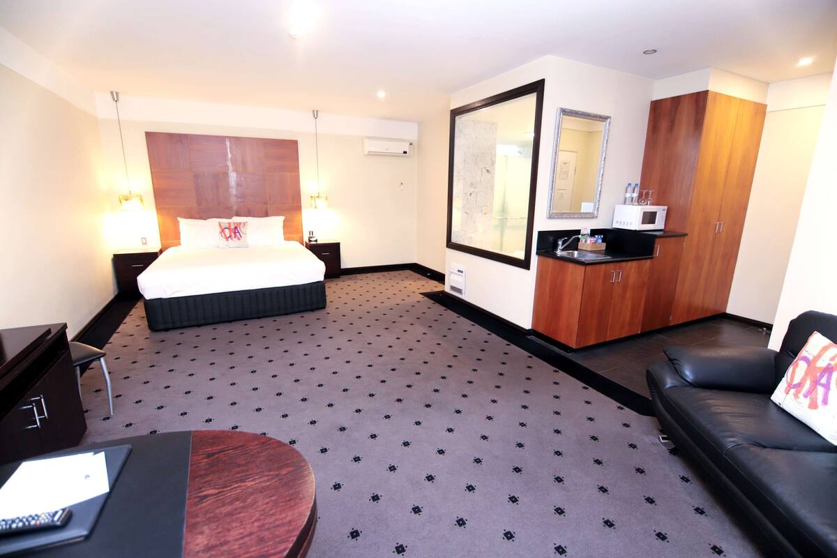 A hotel room with a bed, a desk, a sofa, a mirror, and a kitchenette.