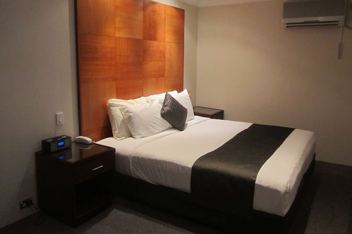 A hotel room with a large bed, a wooden headboard, a bedside table with a phone and a charging device, and an air conditioning unit on the wall.