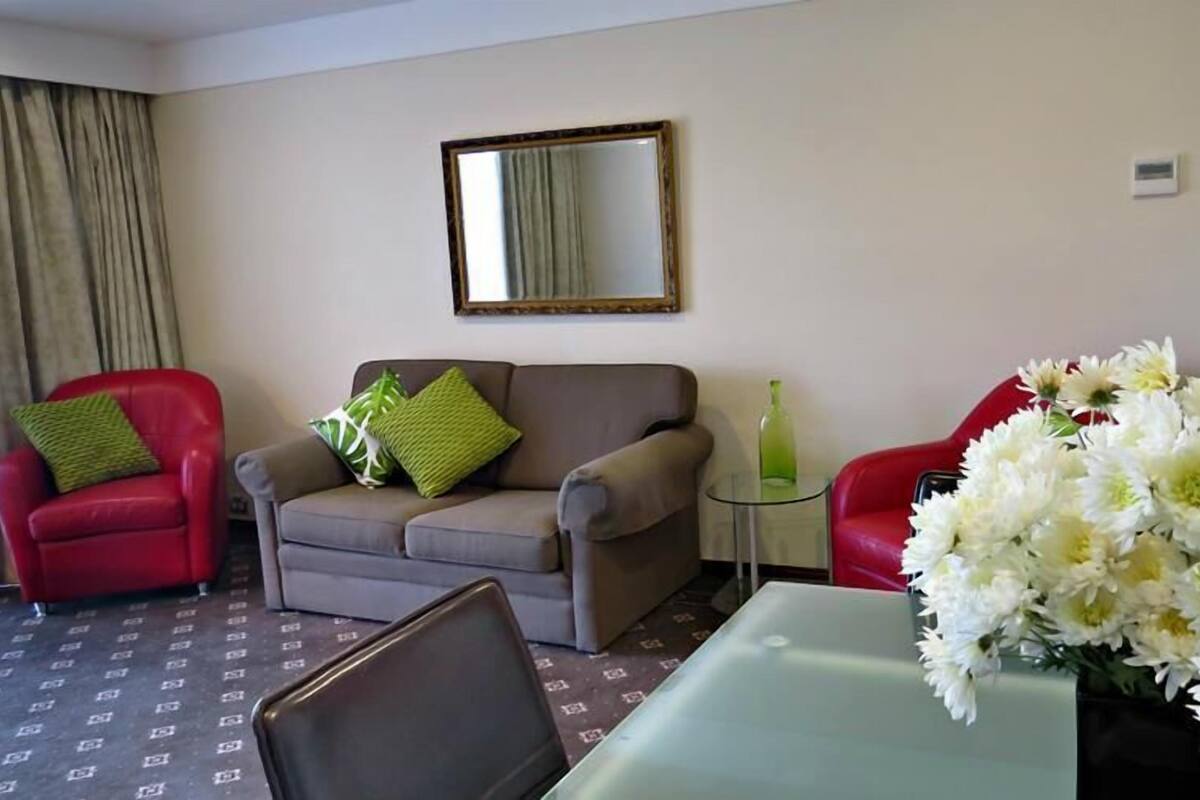 A hotel room with a grey sofa, a red armchair, a small table with a vase of flowers, and a mirror on the wall.