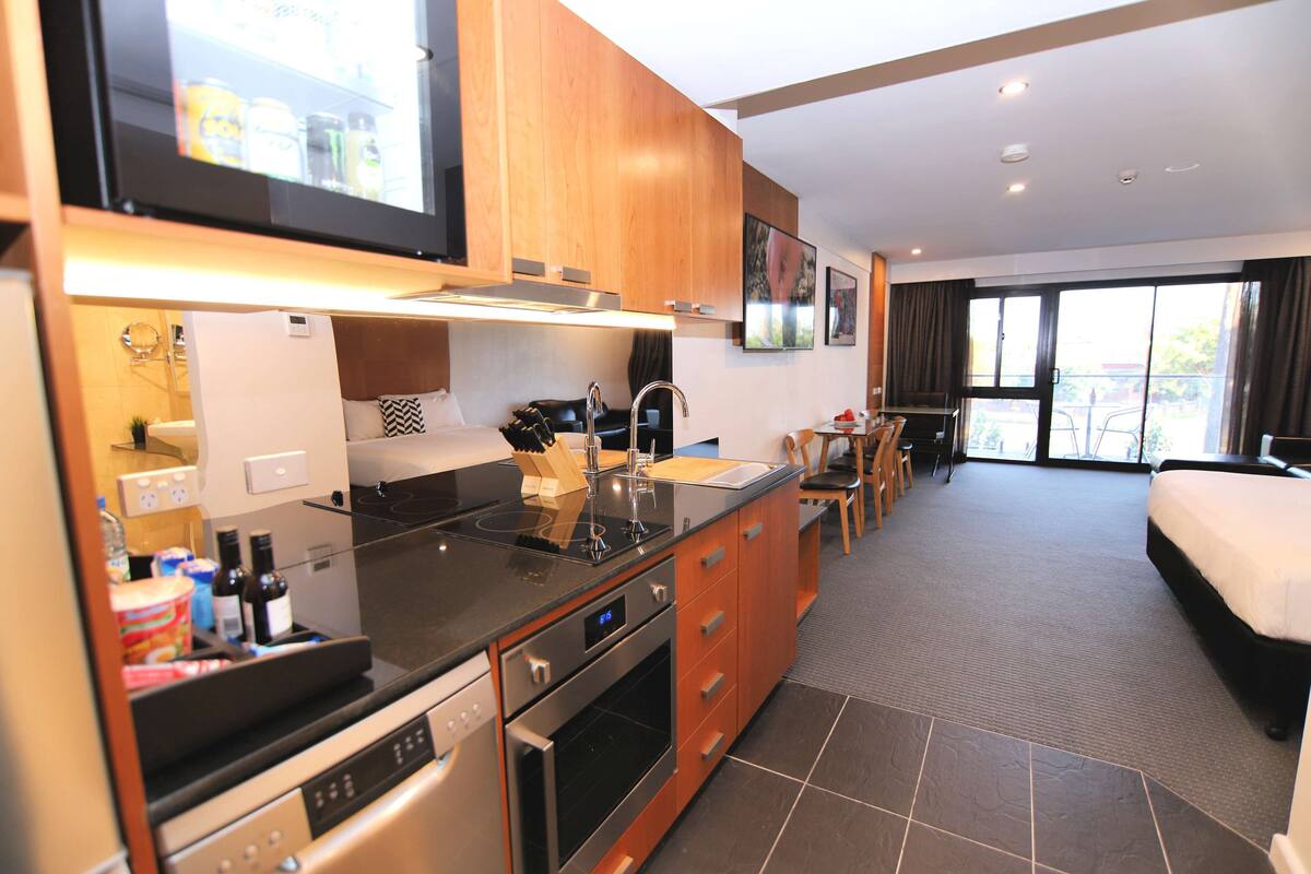 A modern kitchen with a built-in oven, a dishwasher, and a sink. There is a dining area with a table and chairs. The room has a large window and a bed.