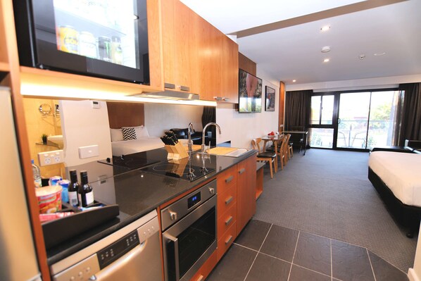 A modern kitchen with a built-in oven, a dishwasher, and a sink. There is a dining area with a table and chairs. The room has a large window and a bed.