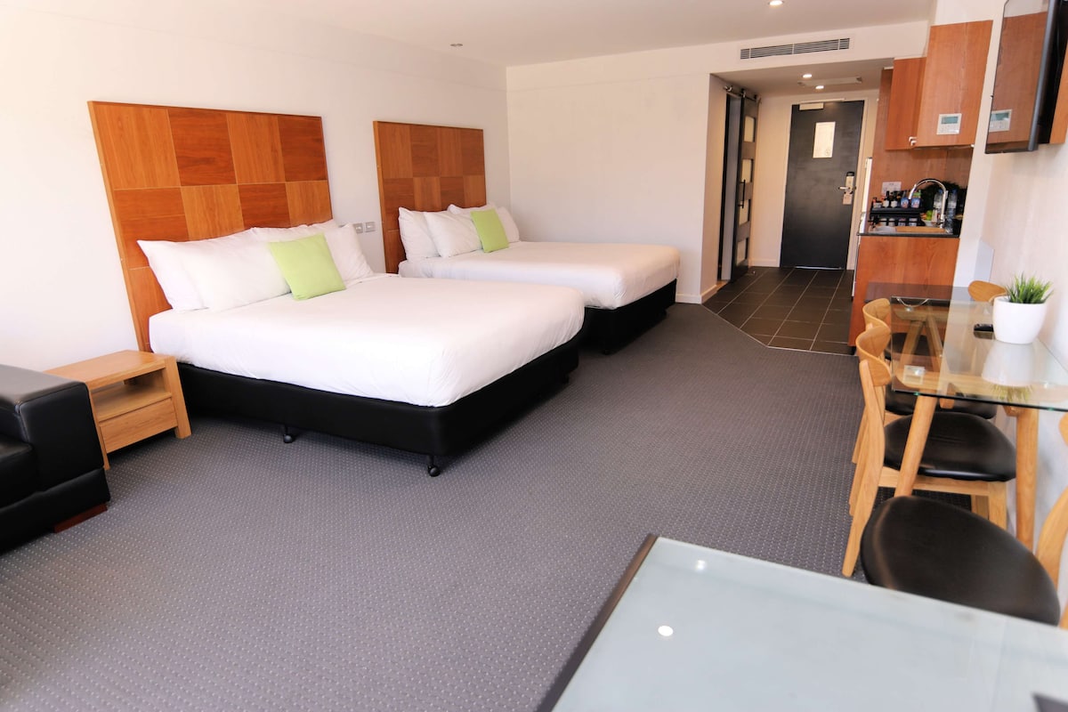 A hotel room with two beds, a desk, a chair, a small table, and a kitchenette area.