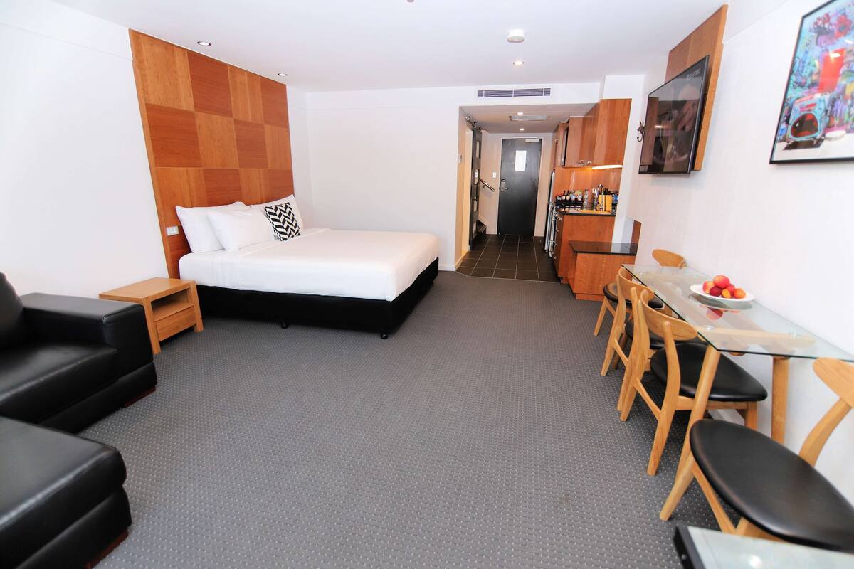 A hotel room with a bed, a small desk with chairs, a kitchenette, and a TV.