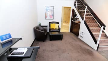 Um saguão de hotel com uma mesa de recepção, uma poltrona de couro, uma pequena mesa com um travesseiro amarelo e uma escada que leva a um banheiro.