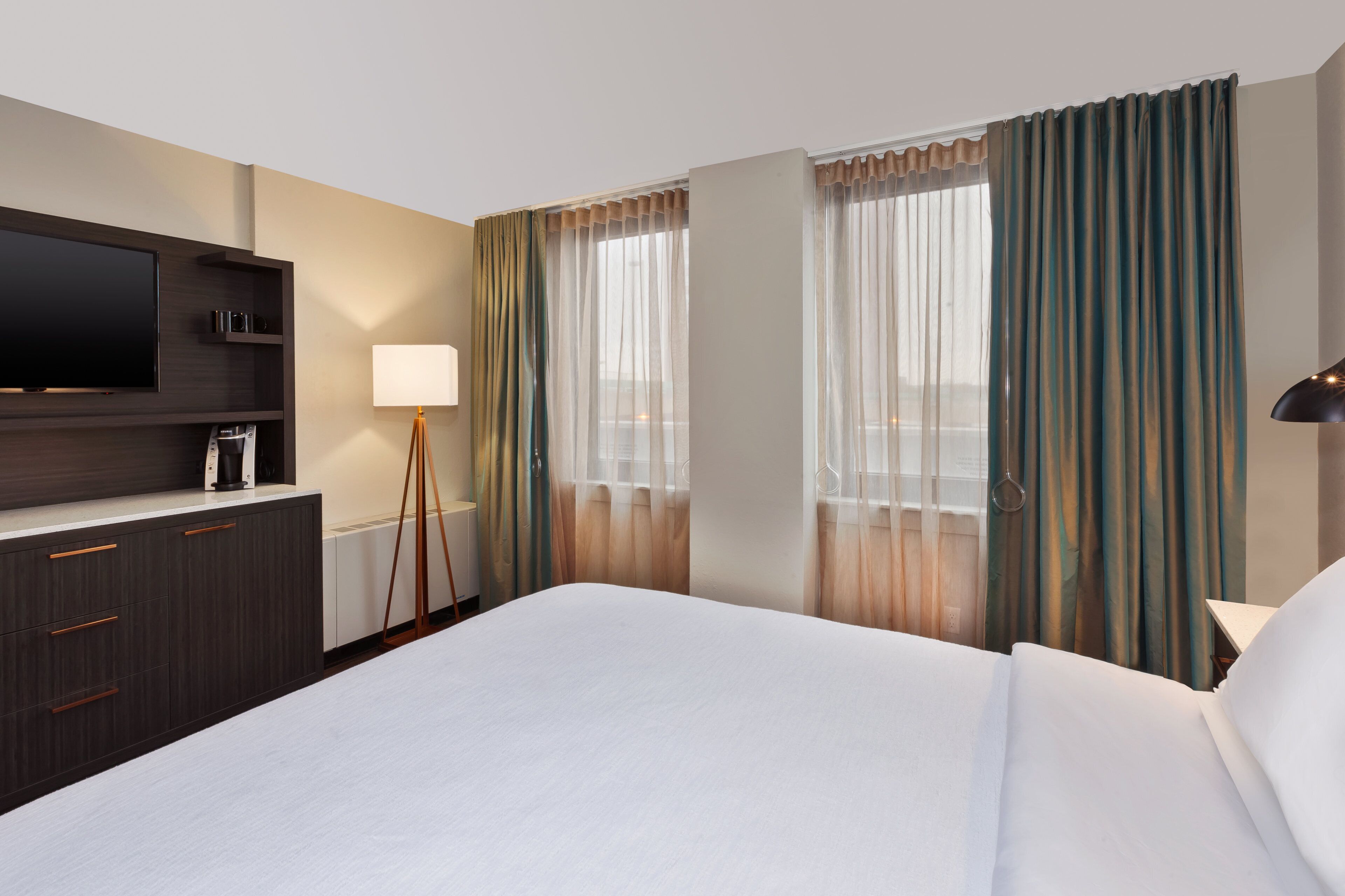 A hotel room with a large bed, a flat-screen TV mounted on the wall, a bedside table with a lamp, and a window with curtains.