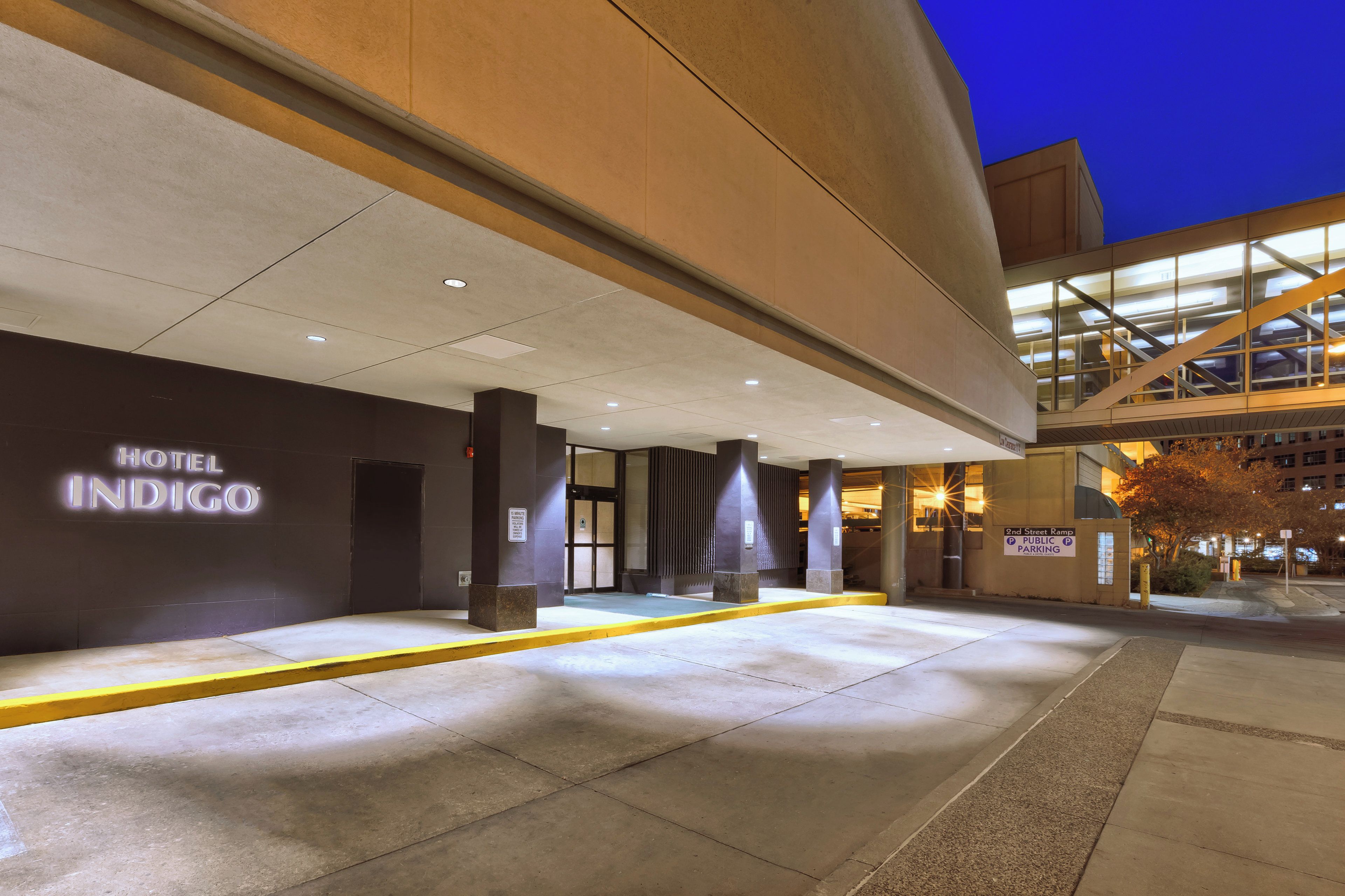 Entrada de hotel con diseño moderno, un letrero que dice 'Hotel Indigo' y una rampa que conduce al estacionamiento.