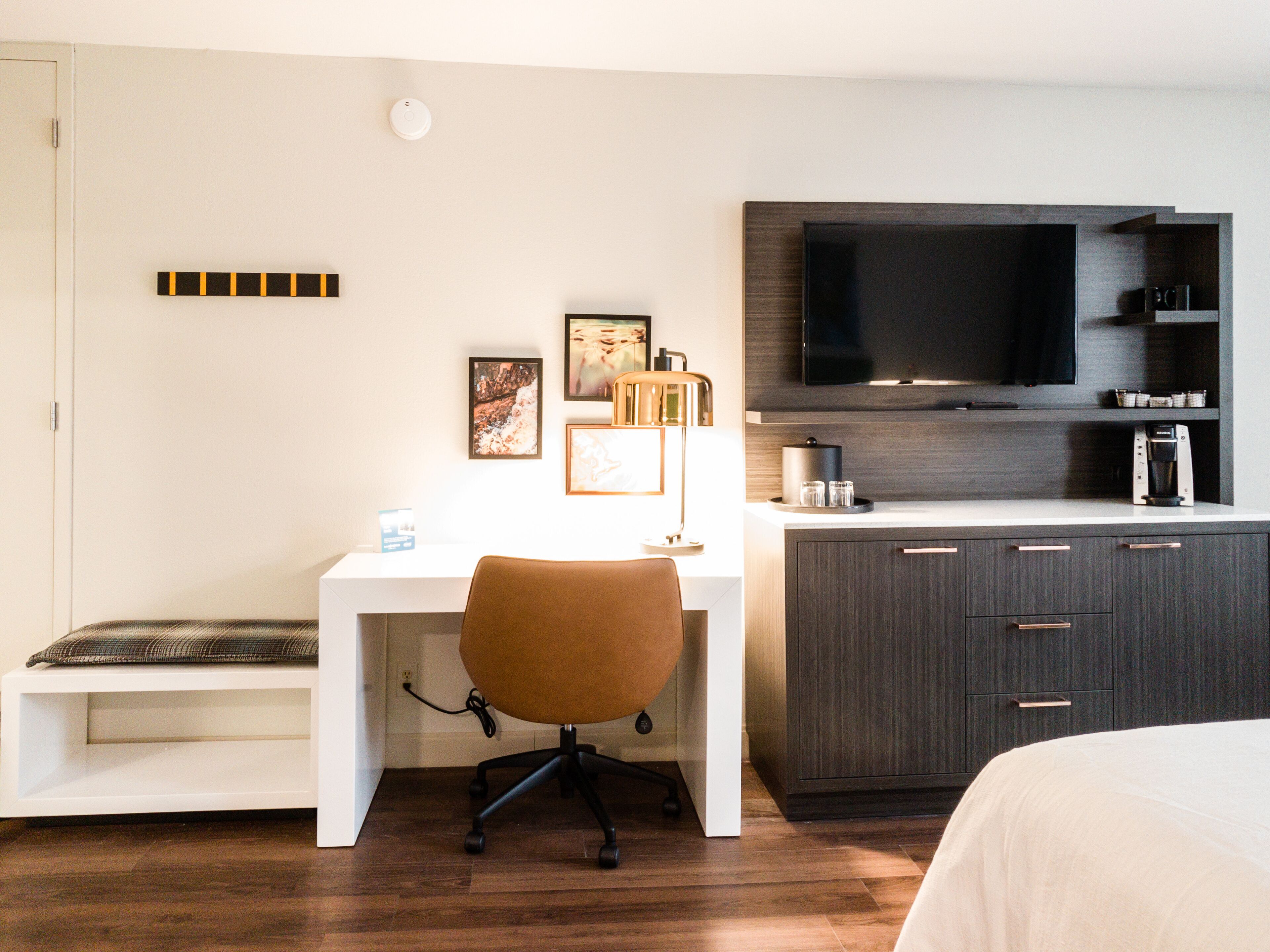 A modern hotel room with a white desk, brown office chair, a flat-screen TV mounted on the wall, a dark wooden cabinet, and a bed with white bedding.