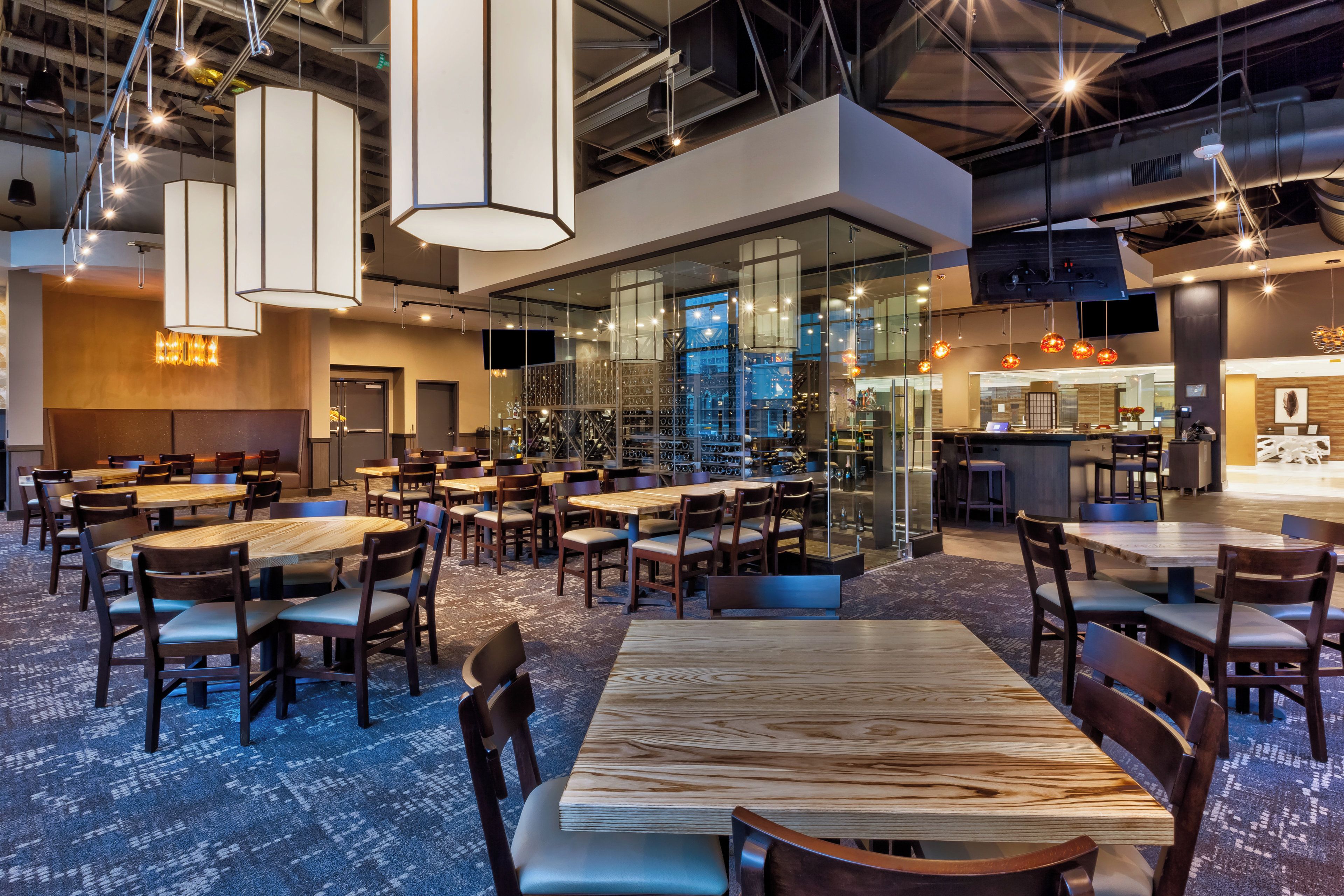 A modern restaurant interior with multiple dining tables and chairs, glass partitions, and hanging pendant lights.