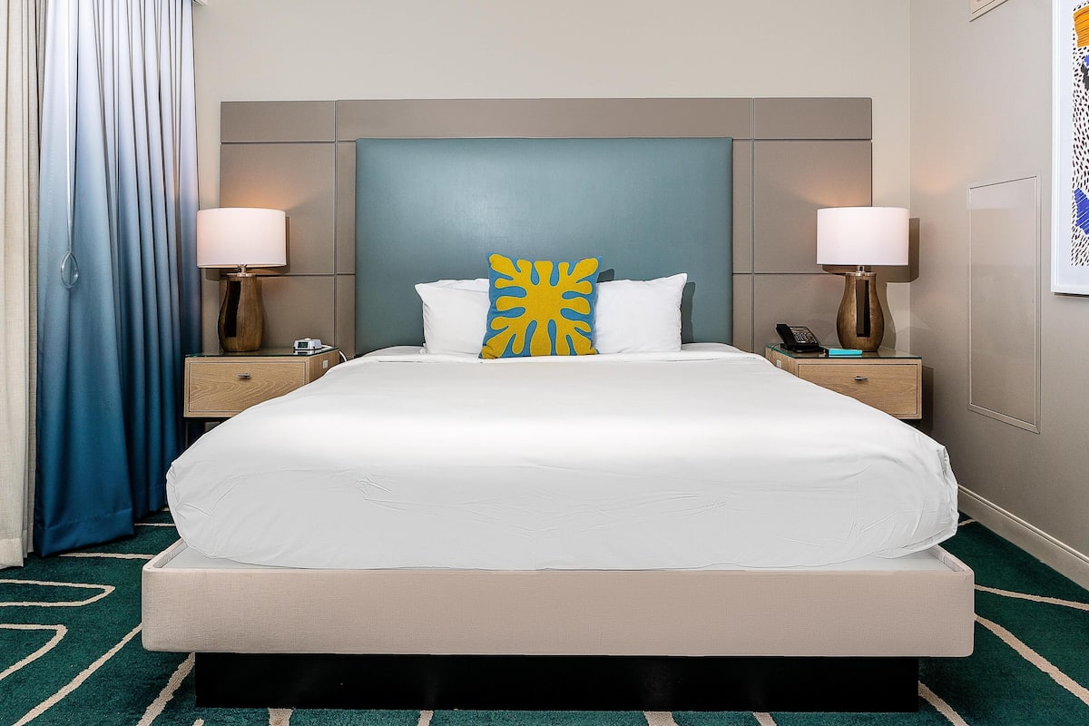 Une chambre d’hôtel avec un grand lit, deux tables de chevet avec des lampes, une petite table et un rideau bleu.