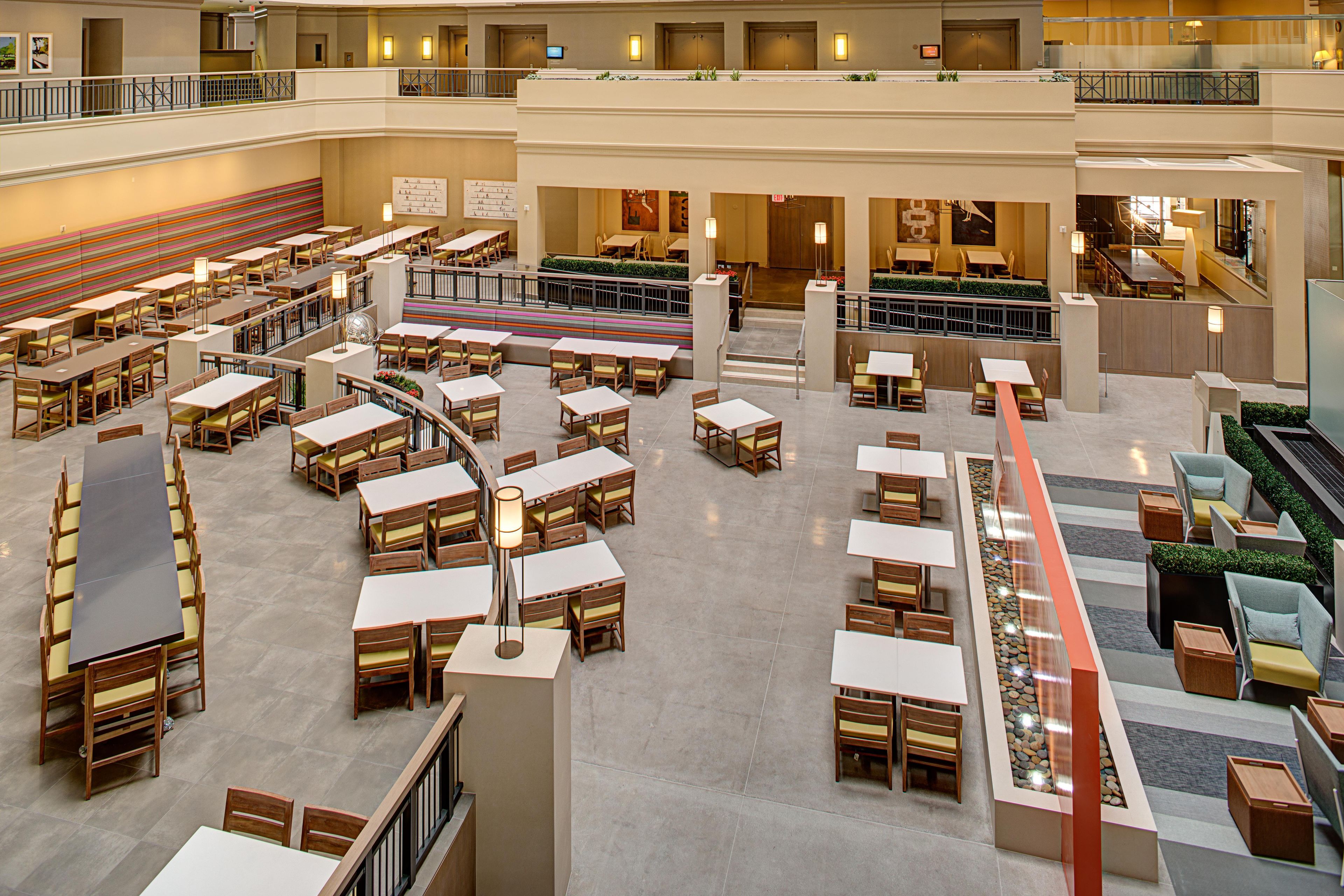 A spacious hotel lobby with multiple dining areas, a central staircase, and a second-floor balcony.