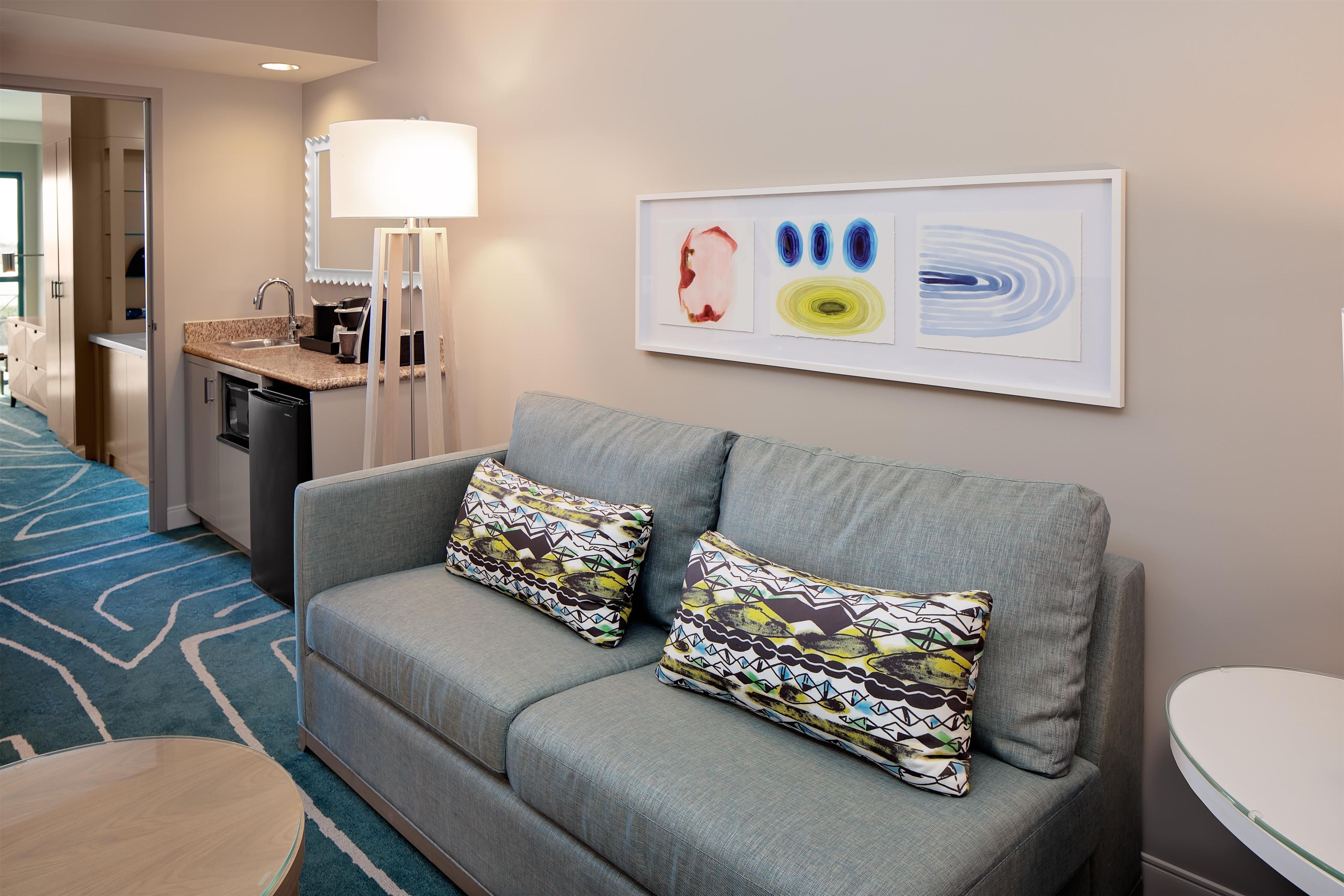A modern hotel room with a grey sofa, patterned cushions, a small round table, a glass-top coffee table, a kitchen area with a sink and a stove, and abstract art on the wall.