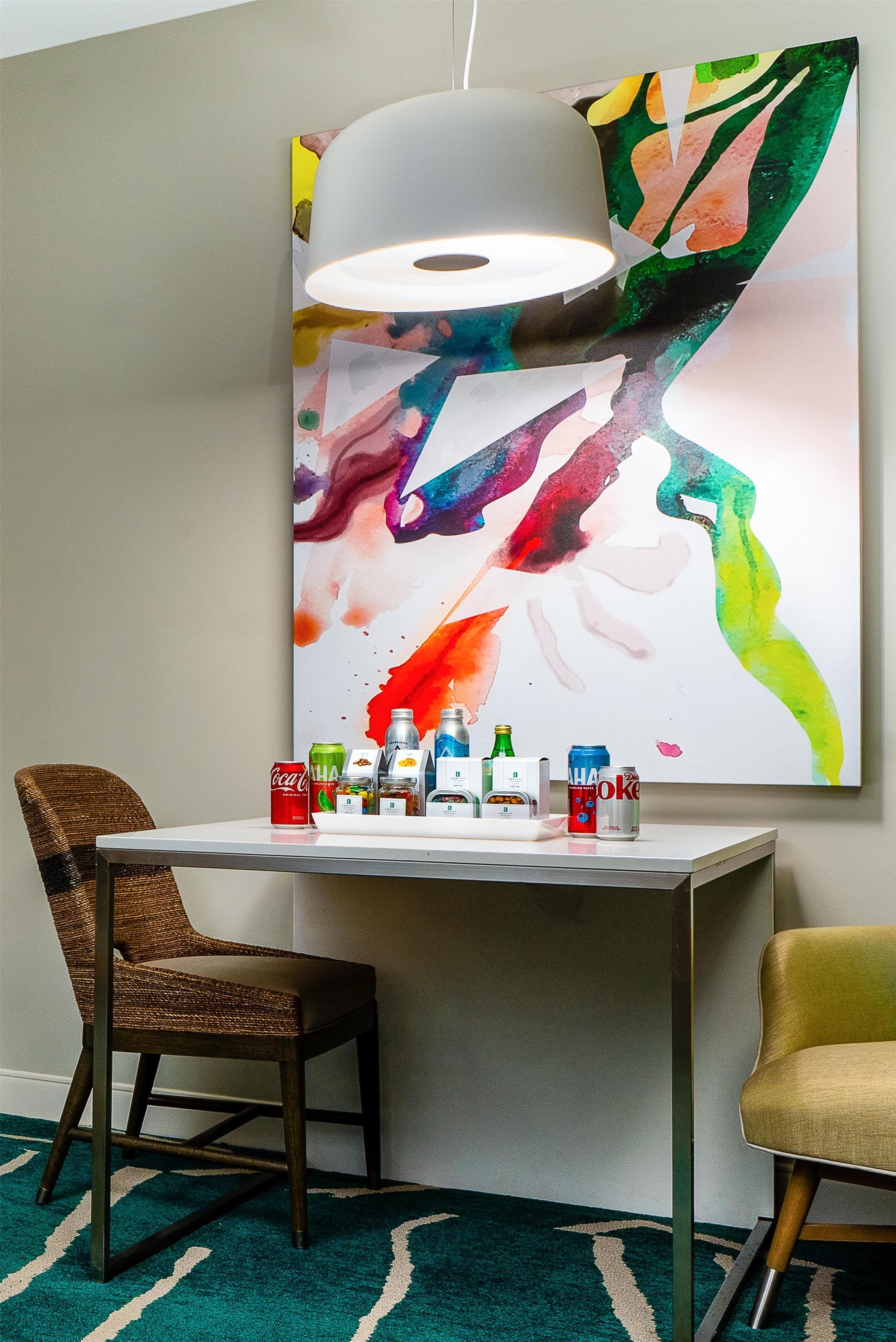 Een moderne kamer met een tafel waarop verschillende drankjes staan, een kleurrijk abstract schilderij en twee stoelen.
