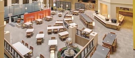 Un espacio interior amplio con varias mesas y sillas para comer, una fuente decorativa central y un balcón en el segundo piso con barandilla.