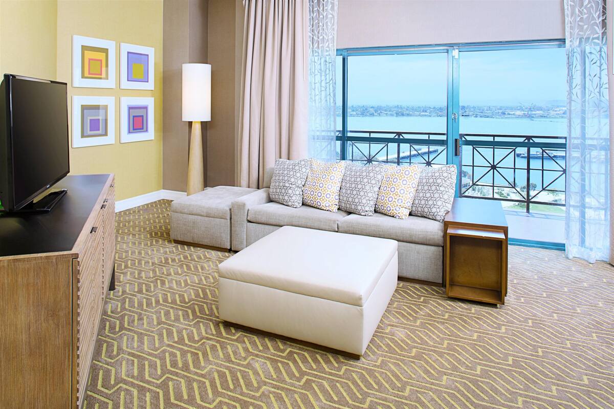 Une chambre d’hôtel équipée d’un canapé, d’un pouf, d’une télévision et offrant une vue sur un plan d’eau depuis un balcon.