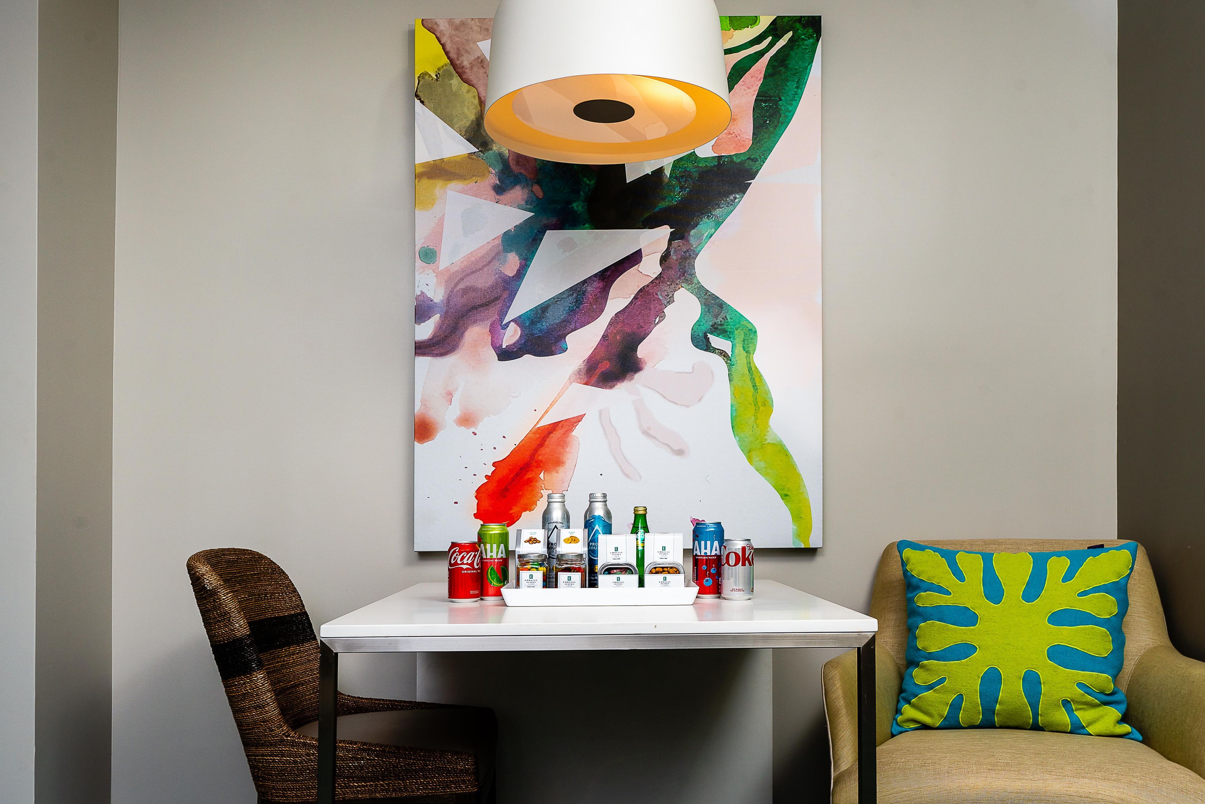 A modern interior with a white table displaying various beverages, a colorful abstract painting, and a chair with a unique pillow.