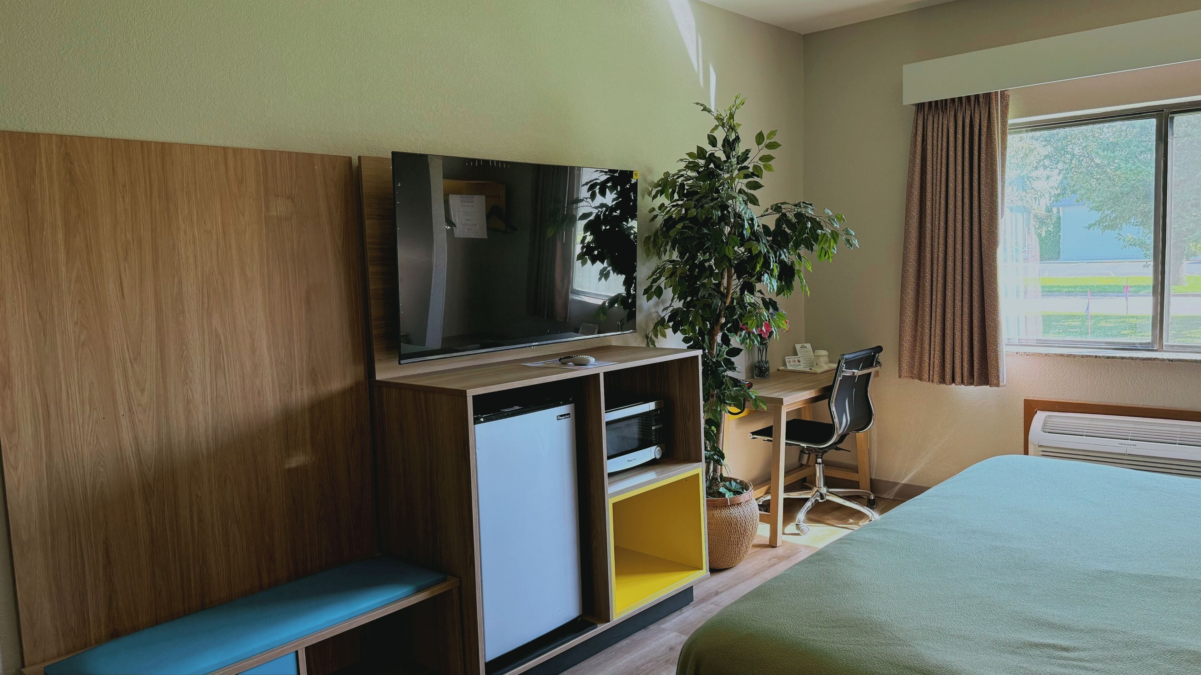 Een hotelkamer met een bed, een bureau met stoel, een televisie, een kleine koelkast en een bloempot.