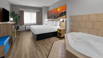 Habitación de hotel con cama, escritorio, televisor, un cuadro, baño con bañera y una planta.