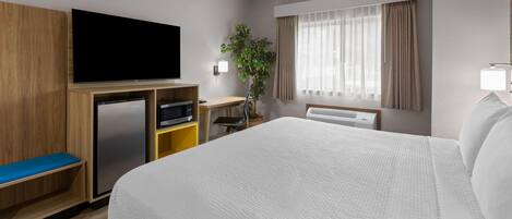 A hotel room with a bed, a television, a desk, a plant, and a window with curtains.