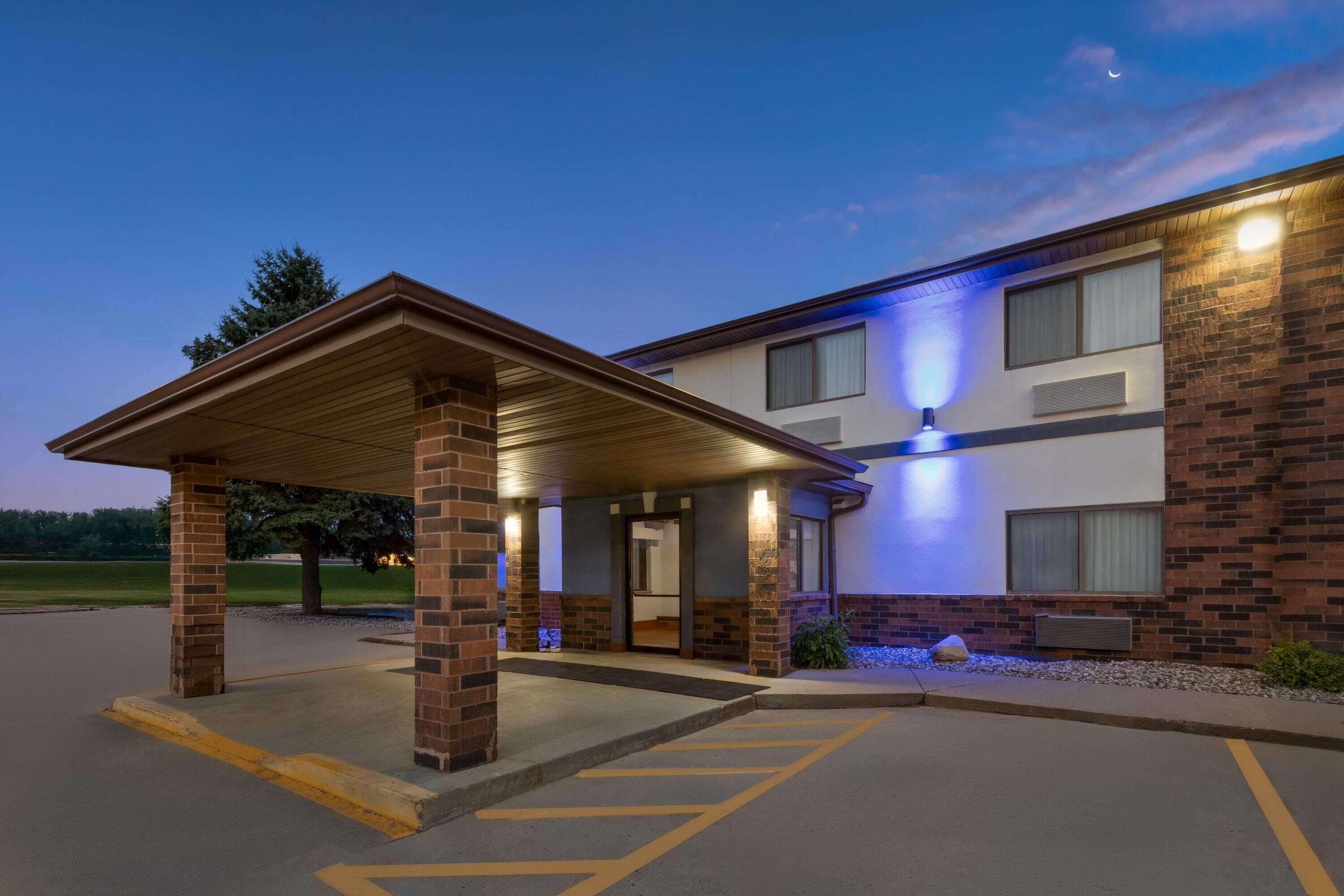 A hotel entrance with a covered waiting area, a parking lot, and a building with a brick facade.
