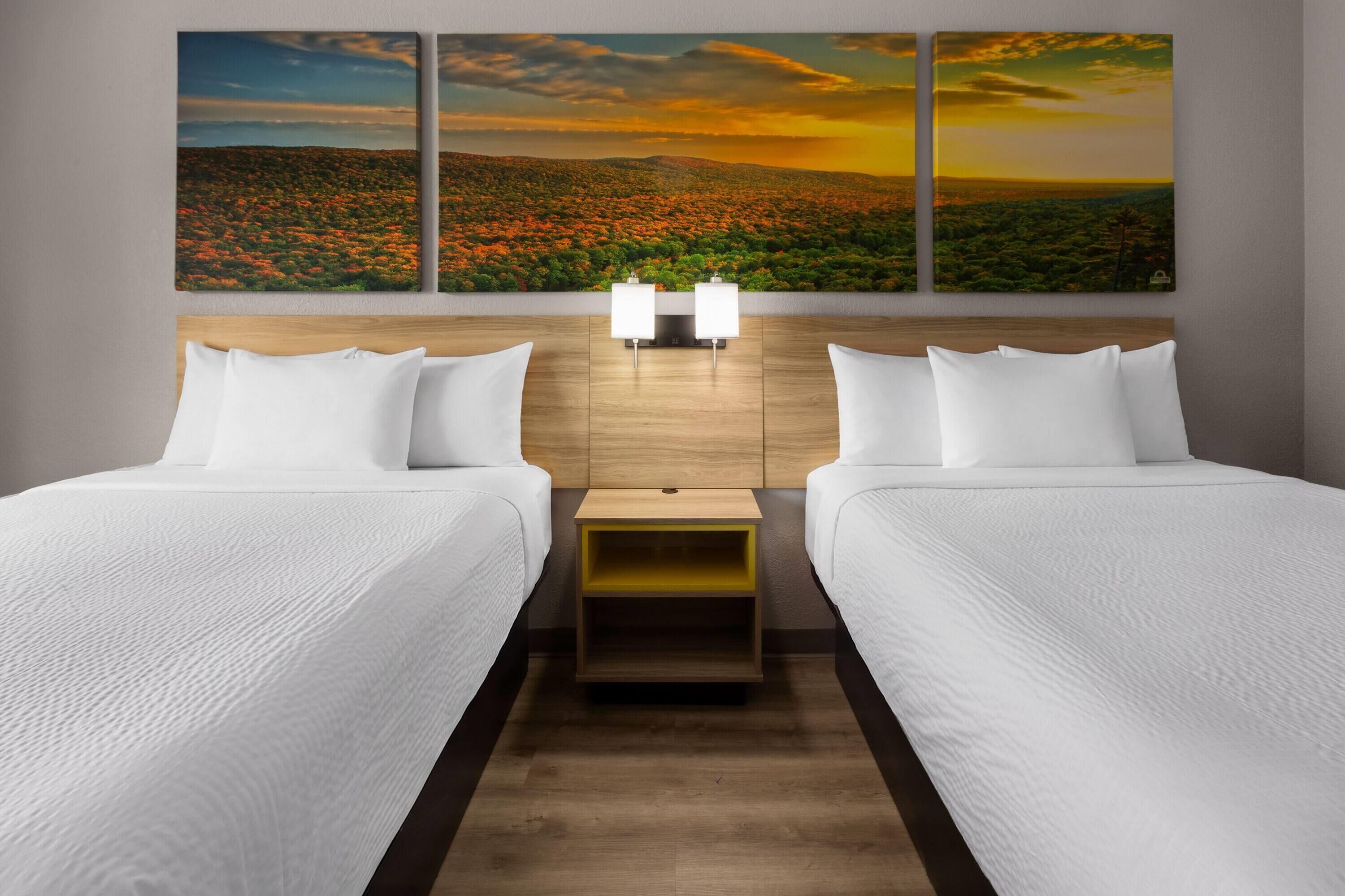 A hotel room with two beds, a wooden headboard, a nightstand with a lamp, and a large landscape painting above the beds.
