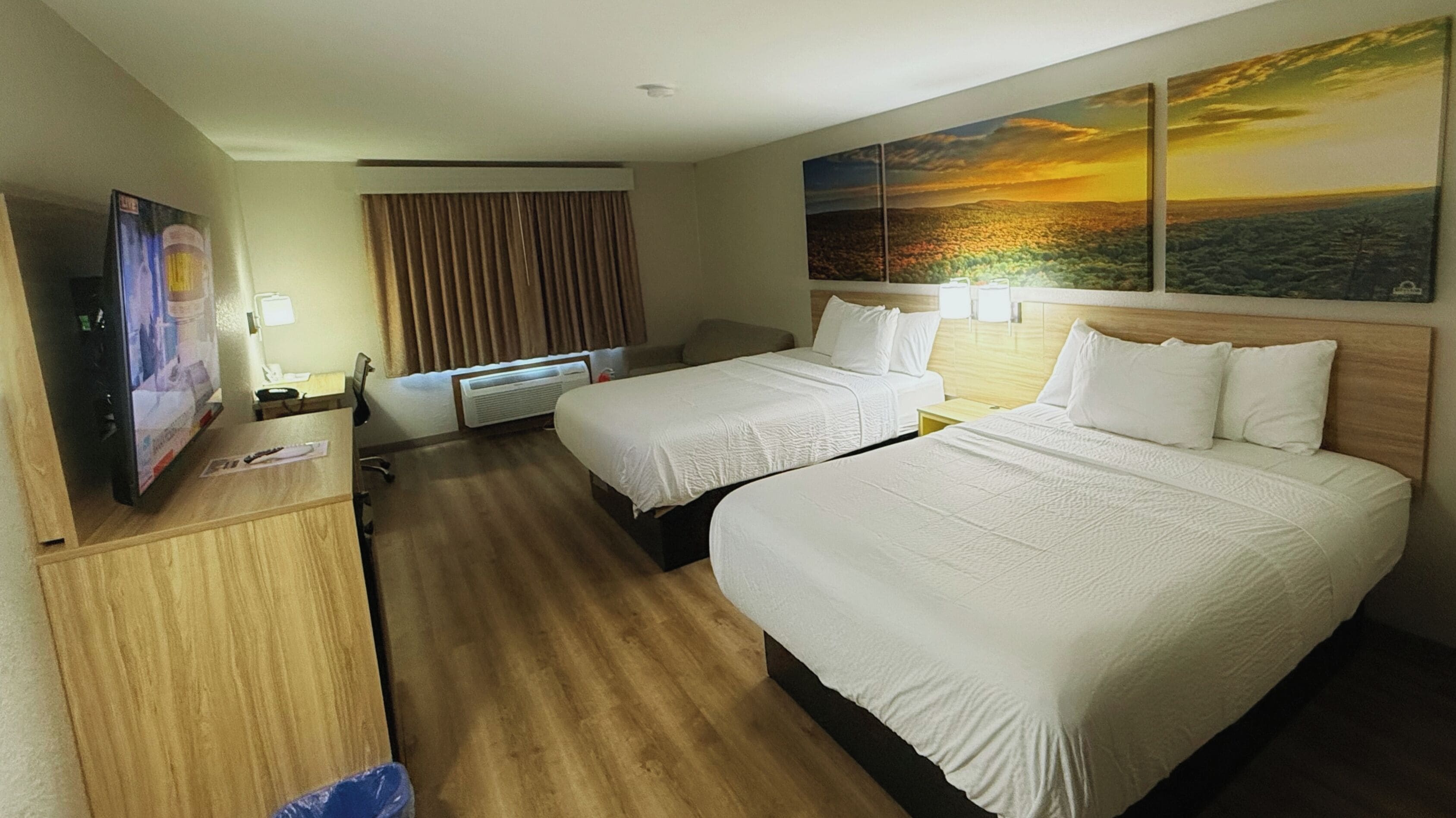 A hotel room with two beds, a desk, a TV, and a painting of a sunset.