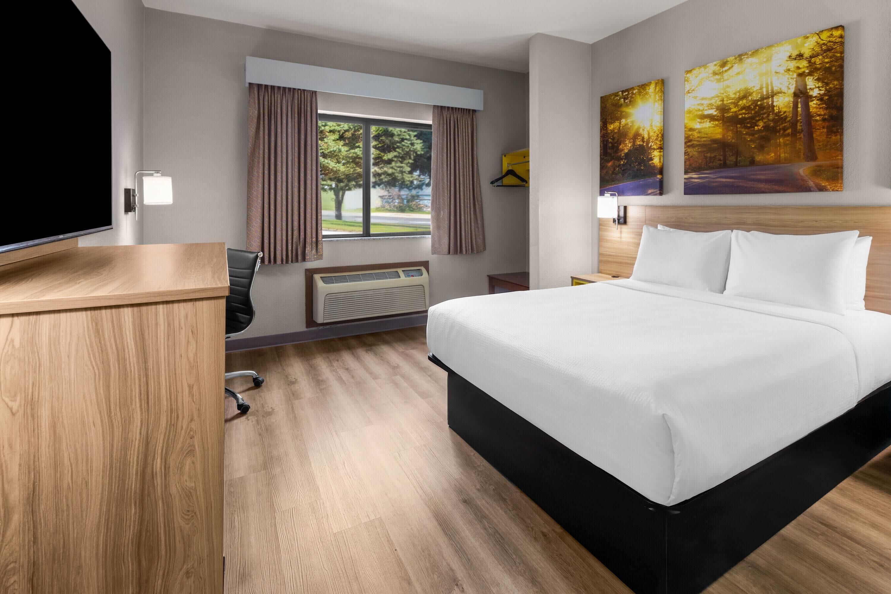 A hotel room with a bed, a desk, a chair, a window with curtains, a wall-mounted TV, and two paintings.