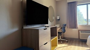 Une chambre d’hôtel équipée d’un téléviseur à écran plat posé sur un meuble en bois, d’un bureau avec une chaise, d’une table de chevet, d’une fenêtre avec des rideaux et d’un appareil de climatisation.