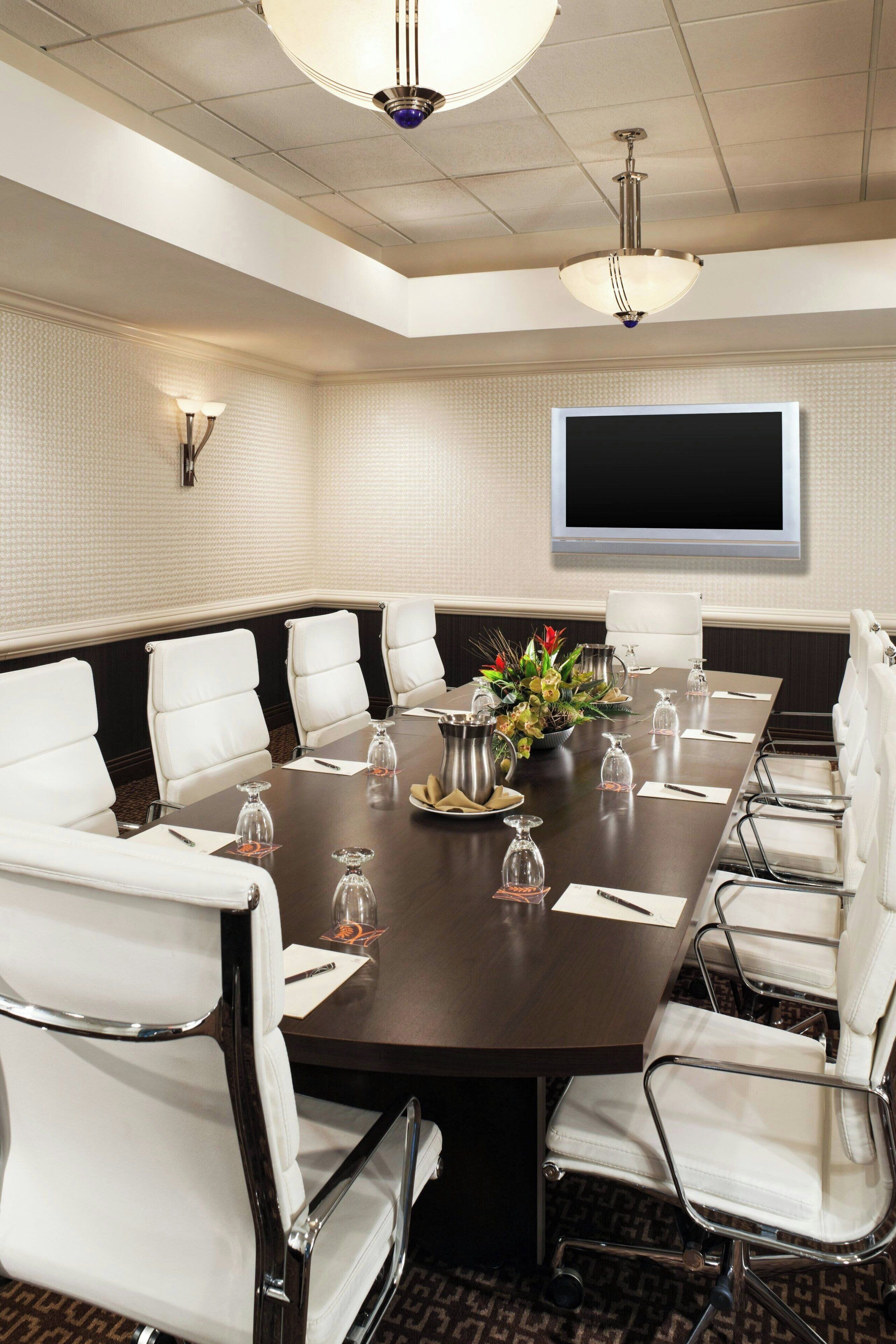 Un salón de conferencias con una larga mesa, sillas de cuero blanco, una pantalla grande y un centro de mesa decorativo.