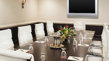 Un salón de conferencias con una larga mesa, sillas de cuero blanco, una pantalla grande y un centro de mesa decorativo.