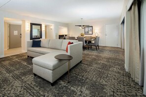 A modern hotel room with a grey sectional sofa, a small round table, a dining area with a table and chairs, and a patterned carpet. - Sheraton Detroit Metro Airport (Romulus)