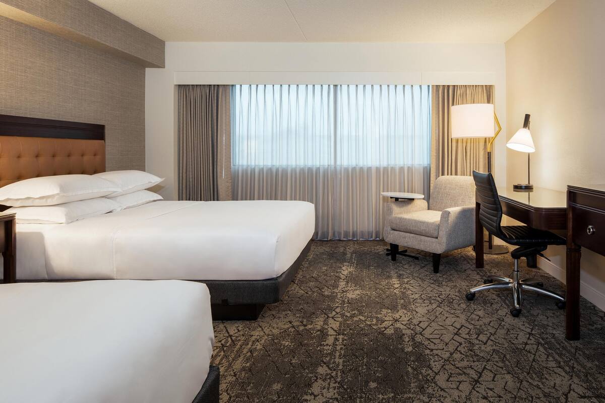 A hotel room with two beds, a desk, a chair, a lamp, and a large window with curtains.