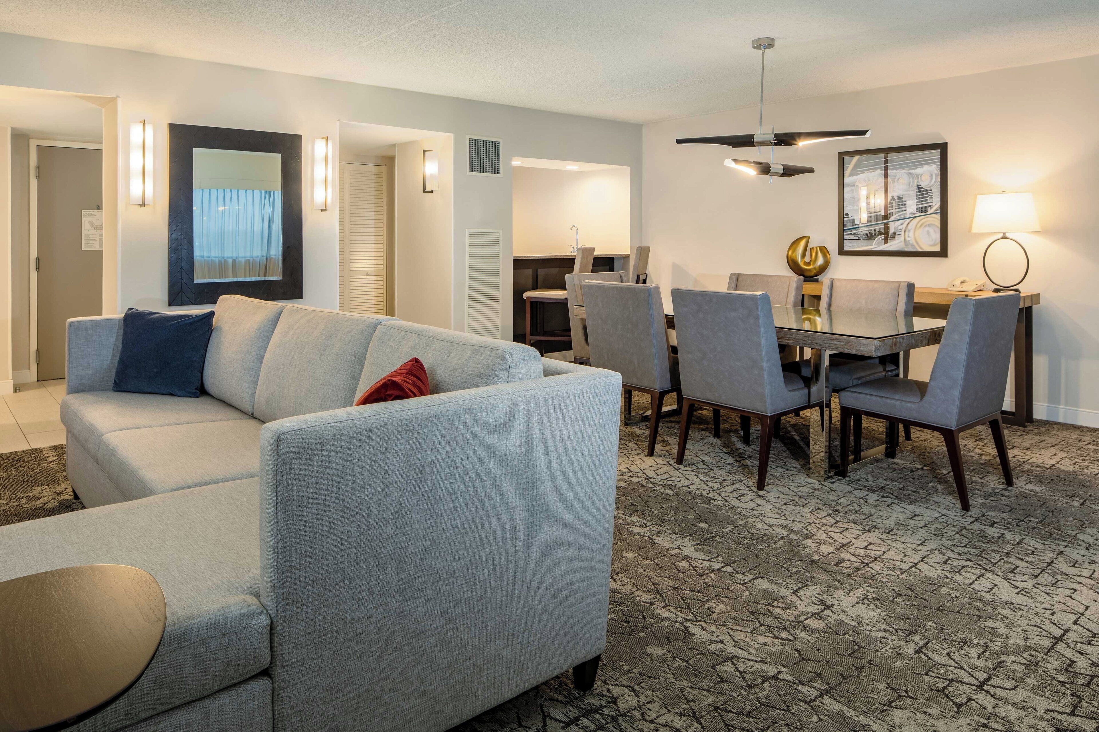A hotel room with a grey sofa, a dining table with chairs, a chandelier, and a cityscape painting on the wall.