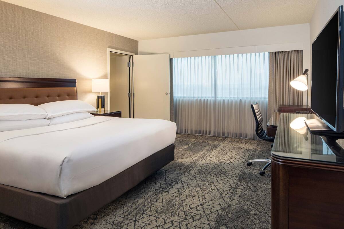 A hotel room with a large bed, a desk with a lamp, a chair, and a TV.