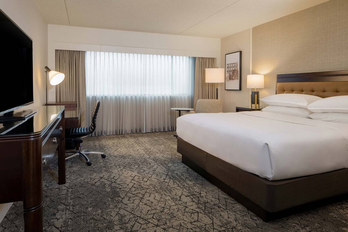 A hotel room with a large bed, a desk with a TV, a chair, a small table, and a lamp.