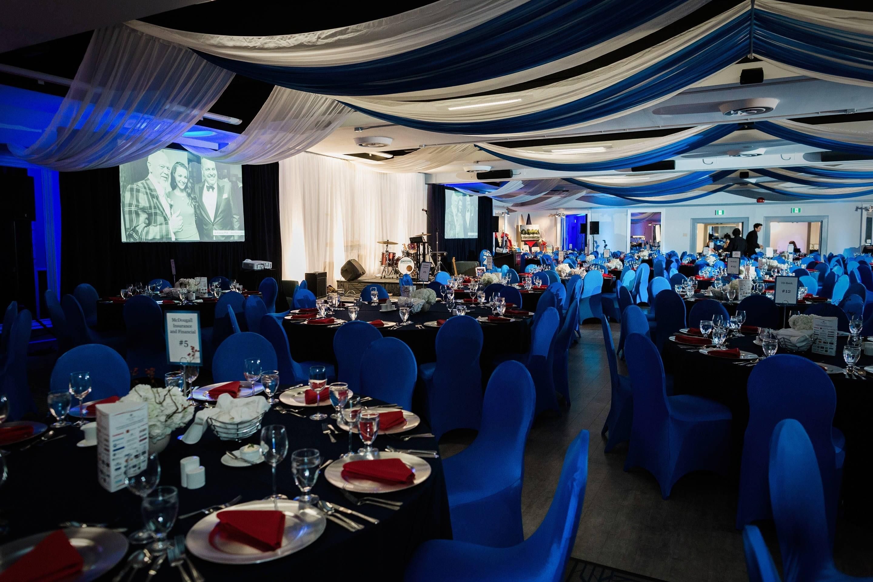 Un salón de banquetes con mesas dispuestas para un evento formal, sillas azules y un escenario con instrumentos musicales.