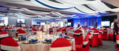 Un salón de banquetes con mesas redondas, cintas rojas y blancas en las sillas y un escenario con una banda.