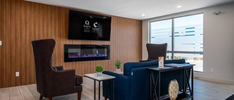 Una sala moderna con una televisión de pantalla plana, un sofá azul, un sillón oscuro, una mesita y un ventanal grande.