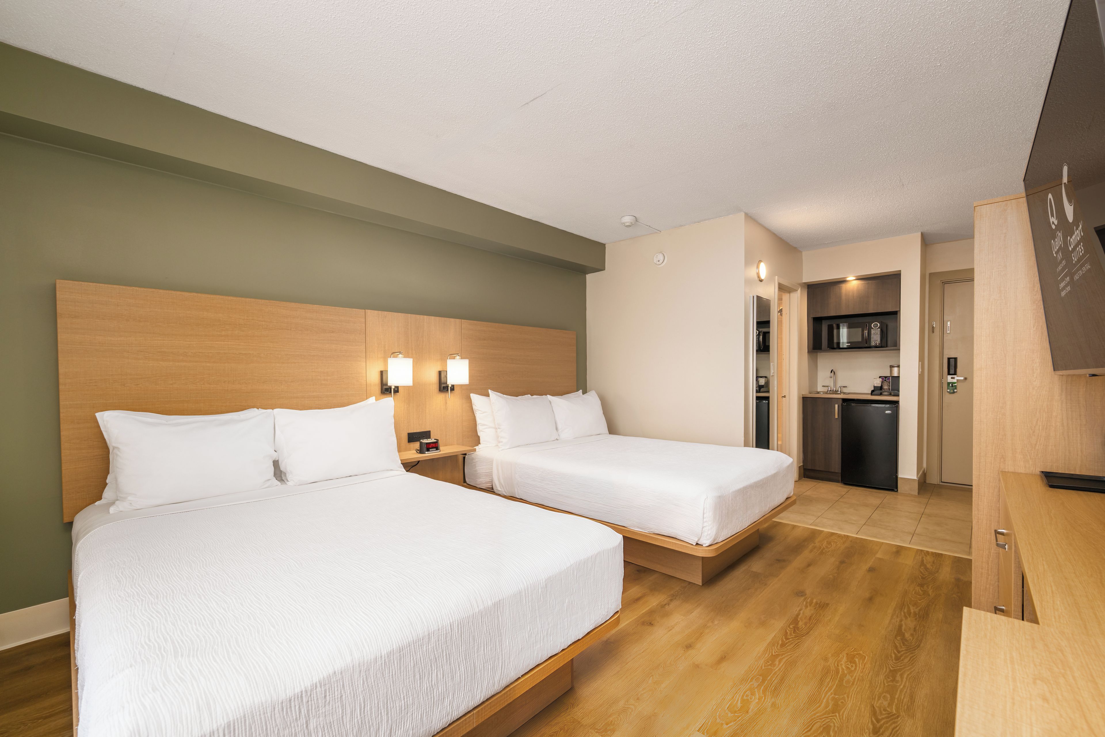 Une chambre d’hôtel avec deux lits, une tête de lit en bois, des lampes de chevet, un petit bureau et une kitchenette équipée d’un four à micro-ondes et d’un réfrigérateur.