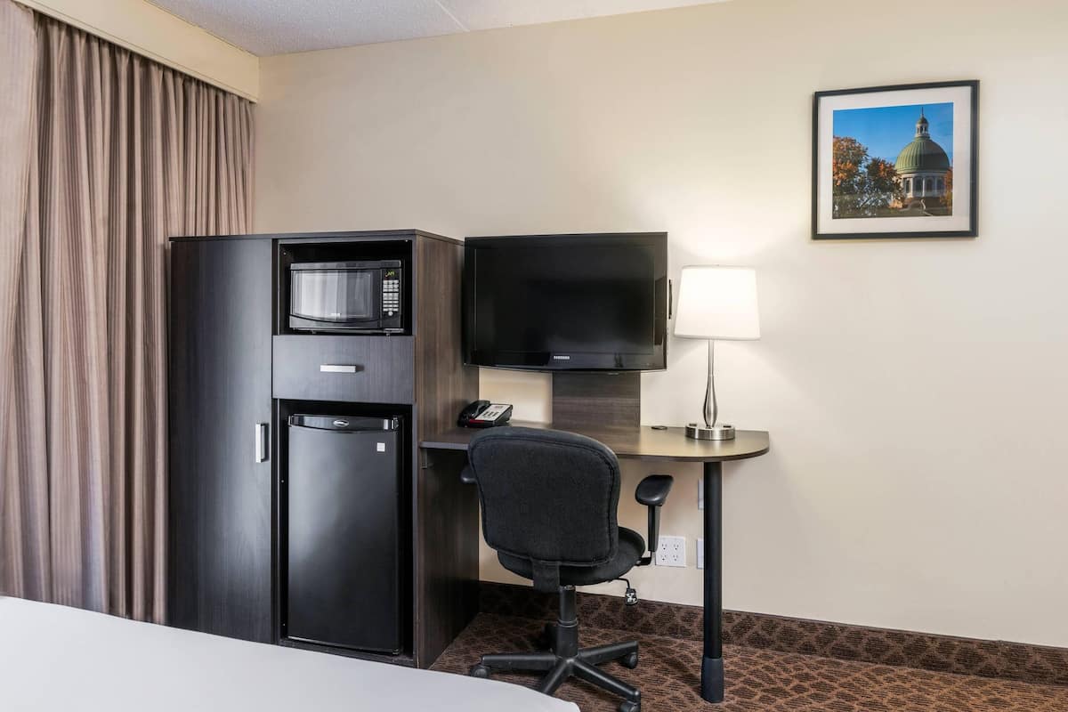 A hotel room with a bed, a desk with a computer, a microwave, a refrigerator, a lamp, and a framed picture on the wall.