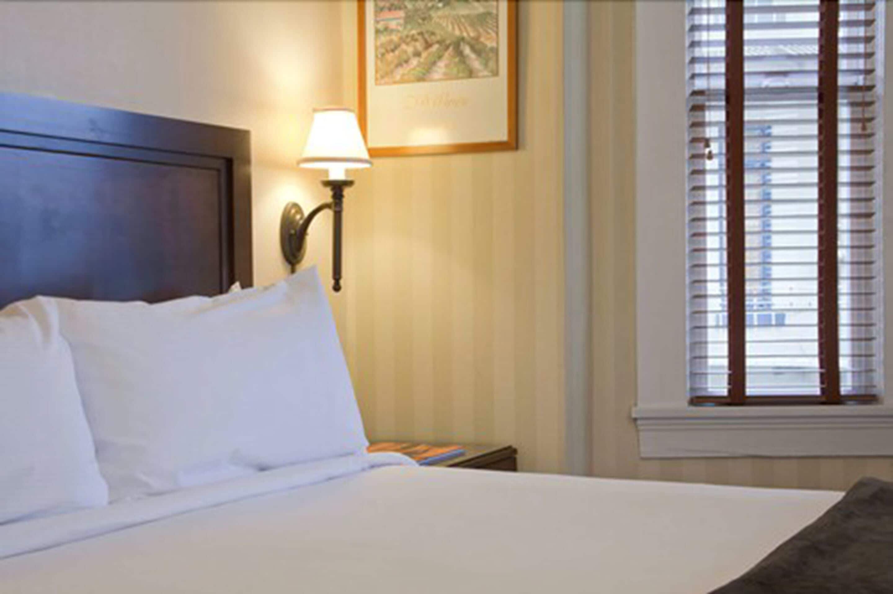 Une chambre d’hôtel avec un lit, une table de chevet, une lampe, un tableau encadré au mur et une fenêtre avec des stores.