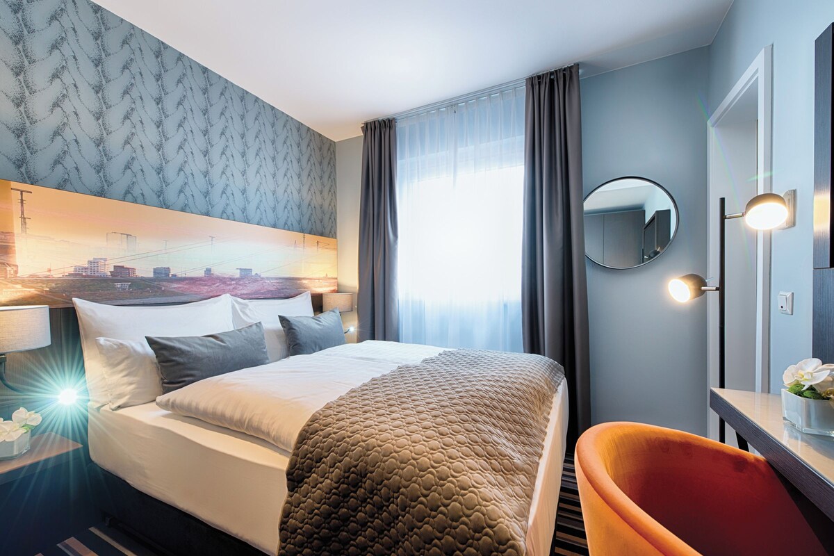 A hotel room with a large bed, a cityscape mural, a round mirror, and a modern chair.