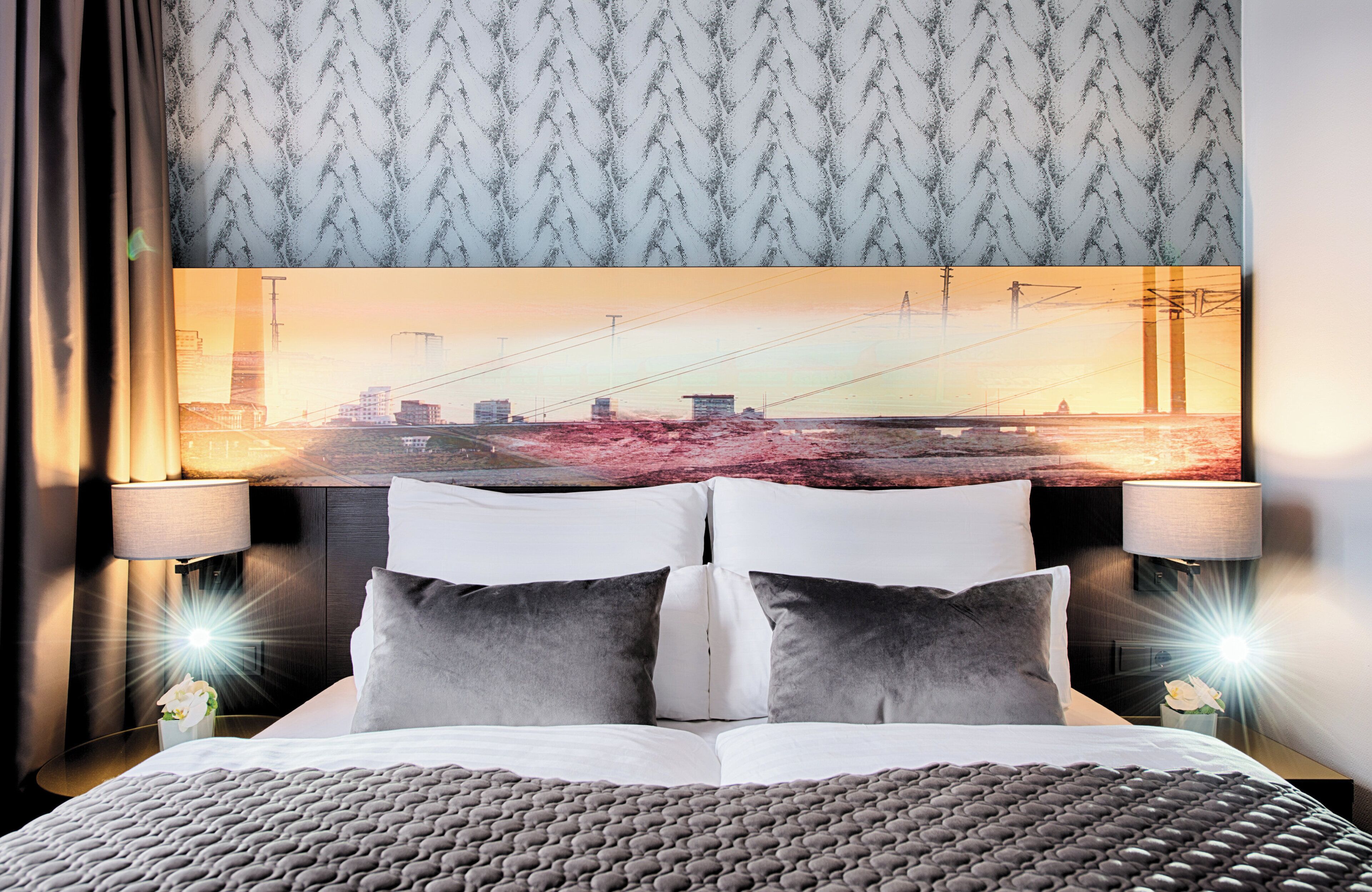 Habitación de hotel con una cama grande, dos lámparas de noche, un cabecero con una pintura de un paisaje urbano y un papel tapiz estampado.