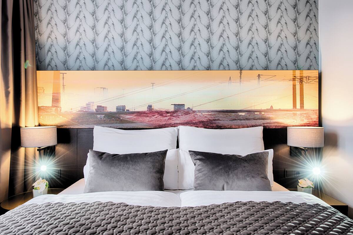 Habitación de hotel con una cama grande, dos lámparas de noche, un cabecero con una pintura de un paisaje urbano y un papel tapiz estampado.