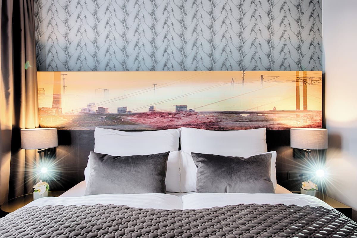 Une chambre d’hôtel avec un grand lit, deux lampes de chevet, une tête de lit ornée d’un tableau représentant un paysage urbain et un papier peint à motifs.
